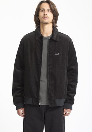 Chaqueta de pana negra con cuello, cremallera frontal y puños acanalados. Presenta un pequeño logo en el lado izquierdo del pecho y un corte holgado.
