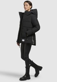 Veste noire matelassée avec capuche, dotée d'une fermeture éclair et d'un bouton-pression, de poches latérales et d'un logo sur le côté. Portée avec des leggings noirs et des bottes.