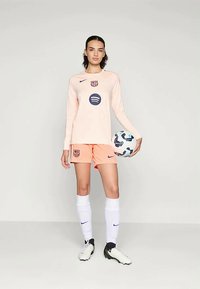 Persikadest piknikärgi jalgpallijersey, millel on FC Barcelona logo, koos persikavärvi lühikeste pükstega, valgete sokkide ja mustade jalgpallijalgadega. Hoides mustrilist jalgpall.