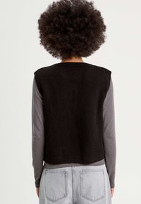 Gilet noir en tricot avec un col rond et des manches courtes, porté sur une chemise à manches longues grise, associé à un jean en denim bleu clair.