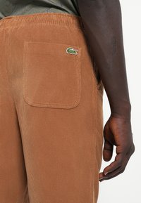 Brown corduroy pants featuring a back pocket and a green and white embroidered crocodile logo. The texture is ribbed and soft.