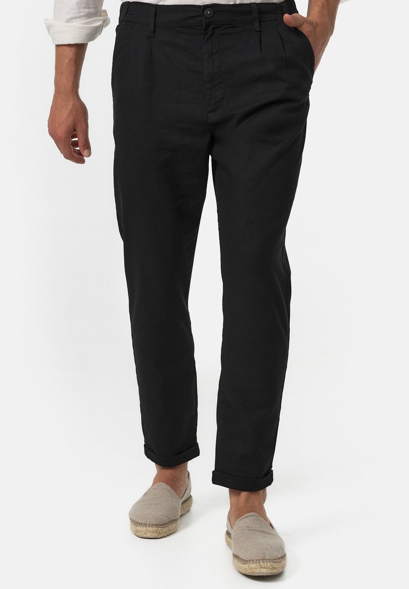 Man in zwart opgerolde broek, beige espadrilles en een wit shirt met opgerolde mouwen, staand met één hand in zijn zak.