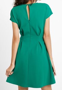 Grön A-linje klänning med korta ärmar, nyckelhål bakstängning och plisserade detaljer vid midjan. Slätt, lätt tyg.