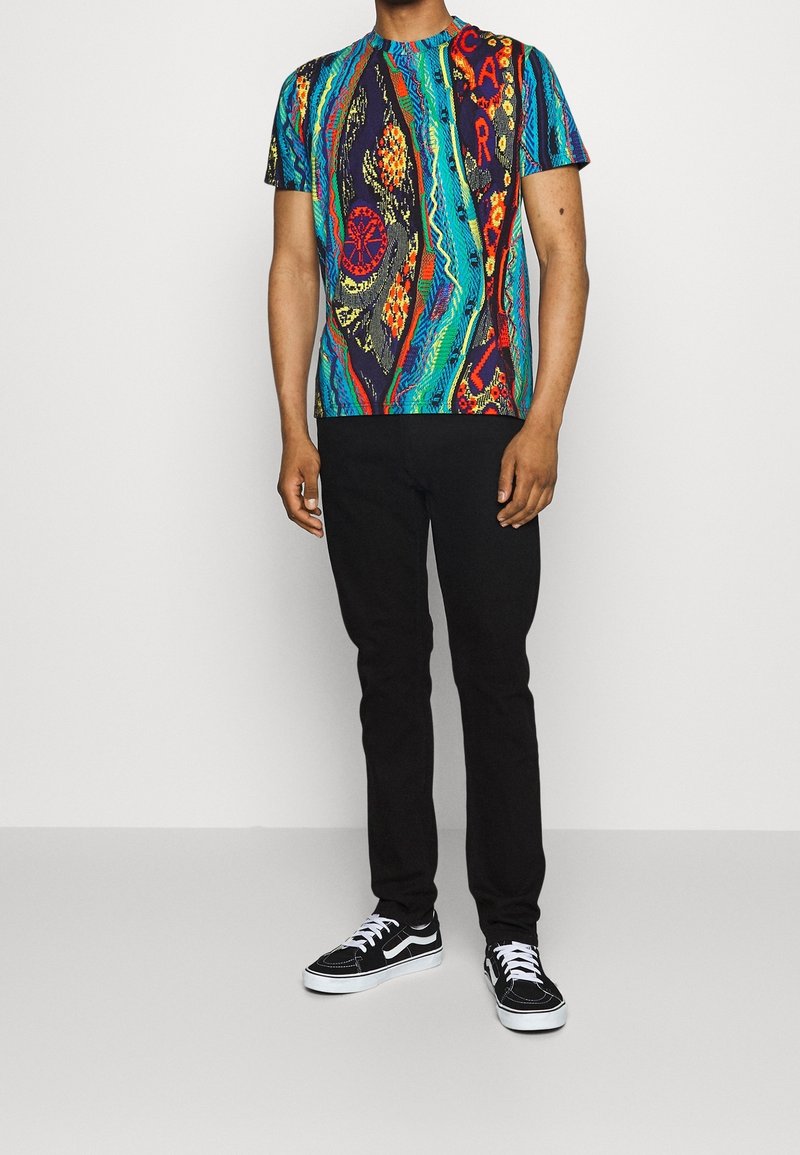 Man med en flerfärgad mönstrad kortärmad skjorta, svarta byxor och svart-vita snörskor som står på ett grått golv.