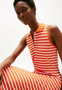 Pomarańczowa sukienka polo bez rękawów z białymi paskami, z kołnierzykiem i guzikami, wykonana z gładkiej tkaniny o luźnym kroju.