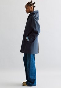 Marine vandtæt frakke med hætte og sidelommer, parret med lyseblå denim og beige fodtøj. Slim fit jeans med brede ben.