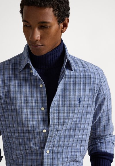 Mėlyna languota medvilninė marškinėliai su sagomis ant apykaklės, kuriuose yra šviesiai ir tamsiai mėlynos spalvos languotų raštų, dėvimi ant tamsiai mėlyno golfo.