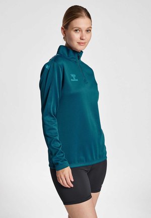 Frau mit blonden Haaren im Pferdeschwanz, die ein türkisfarbenes langärmeliges Viertel-Reißverschluss-Oberteil und schwarze Radsport-Shorts trägt, stehend vor einem schlichten Hintergrund.