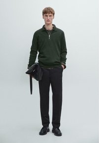 Tmavě zelený svetr na zip, černé kalhoty a černé boty; drží černou tašku. Jednoduchý design, hladká textura a neformální střih.