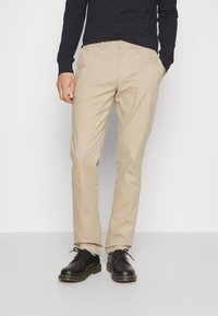 Uomo in pantaloni chino beige, scarpe stringate in pelle nera e una camicia nera a maniche lunghe, in piedi contro uno sfondo bianco.