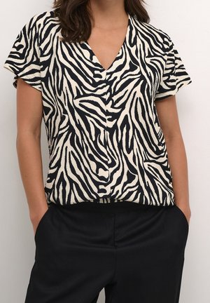 Femme portant un haut à manches courtes et encolure en V avec des rayures zébrées noires et blanches, associée à un pantalon noir, mains dans les poches.