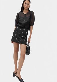 Haut noir transparent à manches bouffantes, boutonné, associé à une mini-jupe noire ajustée ornée de broderies florales argentées. Sac à main noir.