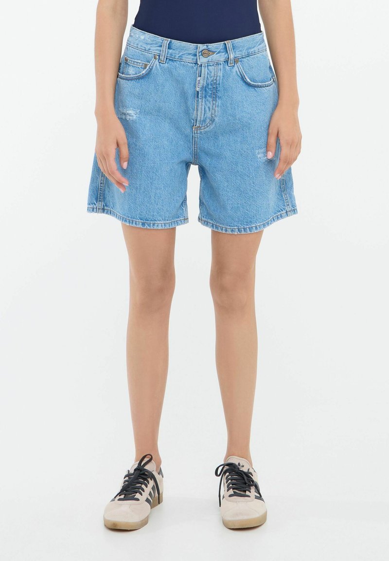 Pantalones cortos de mezclilla azul claro con corte holgado, con bordes deshilachados, cinco bolsillos y pasadores para cinturón. Combinados con zapatillas crema.