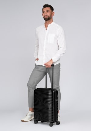 Hartschalentrolley in Schwarz mit geriffeltem Design, kombiniert mit einem leicht strukturierten weißen Hemd und grauen Hosen. Verfügt über einen ausziehbaren Griff und Rollen.