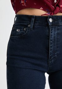 Tmavomodré džínsy s vysokým pásom z denimu. Medzi vlastnosti patrí predné zapínanie na gombík, päť vreciek a malé logo na zadnom vrecku.