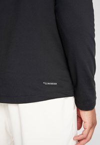 Schwarzes Langarmshirt mit glatter Textur, das ein kleines "CLIMAWARM"-Logo nahe dem Saum trägt, kombiniert mit hellen Hosen.