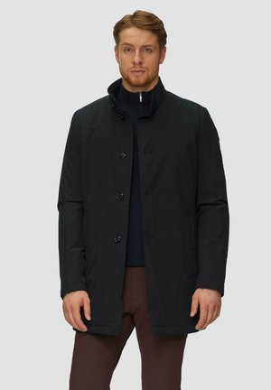 Manteau noir imperméable avec un col haut, fermeture à cinq boutons et poches latérales, porté sur un pull sombre et un pantalon marron. Texture lisse.