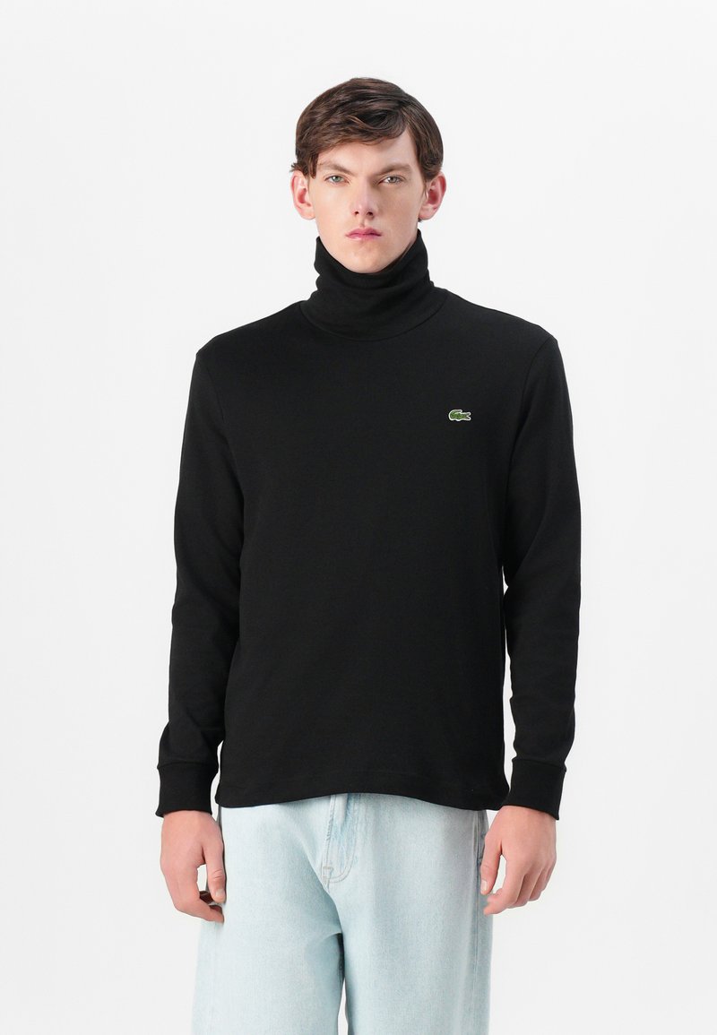 Chandail à col roulé noir en matériau doux et extensible, arborant un petit logo de crocodile vert sur la poitrine gauche. Manches longues, coupe décontractée.