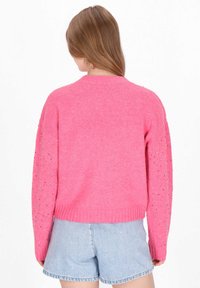 Rózsaszín pulóver puha, texturált felülettel, díszes kivágásokkal az ujjakon és passzos bordázott aljjal. Farmer rövidnadrággal párosítva.