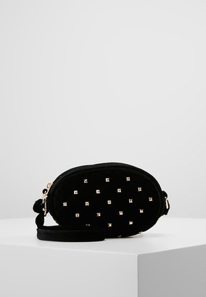 Black oval quilted wristlet bag with gold square studs and zipper closure, placed on a white pedestal against a plain grey background.
