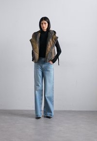 Colete de pele sintética com um padrão texturizado em castanho e dourado, usado sobre um suéter de gola alta preto. Jeans de perna larga em azul claro e sapatos pretos completam o visual.