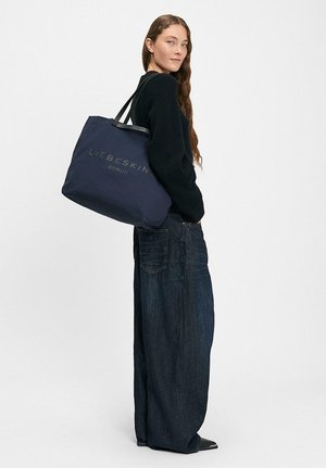 Frau mit langen Haaren trägt schwarzen Pullover und weite dunkle Jeans, sie trägt eine große marineblaue Einkaufstasche mit der Aufschrift "LIEBESKIND BERLIN" über der Schulter.