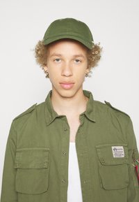 Olive green cap and matching button-up shirt with patch details, large pockets, and a metal zipper accent. Smooth cotton texture.