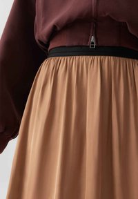 Blusa marrone con zip e maniche lunghe, abbinata a una gonna beige fluida con una vita elastica nera e una texture a pieghe.