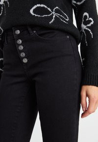 Gros plan d'une personne portant un jean noir taille haute avec quatre boutons métalliques visibles et un pull noir avec des motifs de cœur et de nœud blancs.