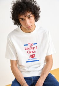 T-shirt branca de algodão com texto em vermelho e azul: "A Escolha Inteligente" e logo "new balance". O modelo tem cabelo encaracolado e está sentado.