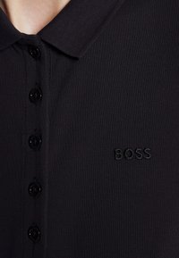 Gros plan d'un polo noir avec cinq boutons noirs et le logo BOSS brodé sur la poitrine.