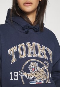 Námořnický modrý mikina s kapucí s předním potiskem, kde je slovo "TOMMY" velkými béžovými písmeny a ilustrací řvoucího tygra, s vrstvenou texturou.