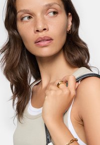 Débardeur beige avec bord blanc, mettant en valeur une bague dorée à la main. Le mannequin a des cheveux bruns ondulés et tient un sac avec une sangle noire.