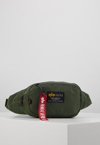 Borsa in nylon verde oliva con tasca frontale con zip, tracolla regolabile e etichetta rossa con il testo "RIMUOVERE PRIMA DEL VOLO". Dettaglio logo in patch.
