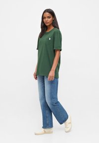 Groene katoenen T-shirt met korte mouwen en een borstzak, gecombineerd met lichtblauwe rechte jeans en beige sneakers.