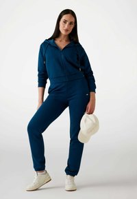 Felpa blu con zip e cordini, abbinata a joggers coordinati, realizzati in tessuto morbido. Indossata con sneakers bianche e un cappellino di colore chiaro.