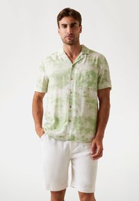 Kurzarmhemd aus hellgrünem Stoff mit Palmenmuster, Holzknöpfen und einer Brusttasche, kombiniert mit weißen Shorts.
