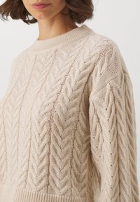 Beige kabelstickad tröja med hög krage, med ett strukturerat mönster och lös passform. Mjukt, lätt stickat tyg som ger en avslappnad look.