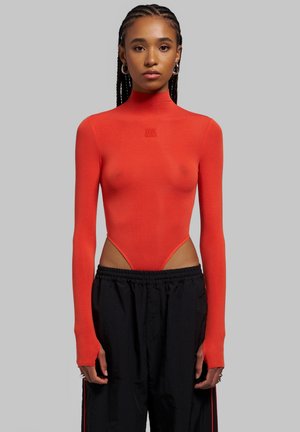 Joven mujer con el cabello trenzado que lleva una camiseta ajustada roja de manga larga con cuello alto y pantalones negros sueltos sobre un fondo gris.