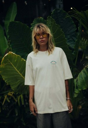 Biała, oversize'owa koszulka z krótkim rękawem, z turkusowym nadrukiem na piersi. Całość dopełniają paskowane spodnie i żółte okulary przeciwsłoneczne.