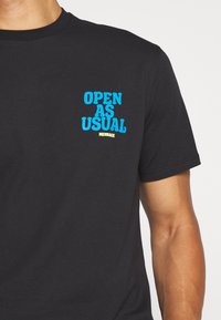 Svart bomull T-shirt med "ÖPPET SOM VANLIGT" i blå och gul grafisk text på vänster bröstområde. Korta ärmar, klassisk passform.