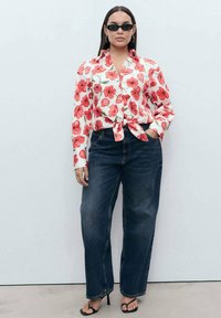 Blusa bianca a maniche lunghe con motivo floreale rosso, legata in vita, abbinata a jeans a vita alta blu scuro e tacchi neri.