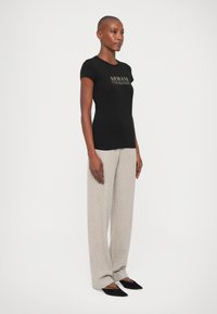 Tricou negru, mulat, cu logo-ul auriu "Armani Exchange", asociat cu pantaloni largi gri deschis și balerini negri cu bot ascuțit. Texturi de țesături fine.