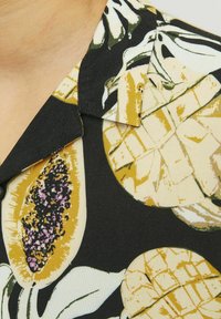 Košulja kratkih rukava od crnog materijala s velikim printom tropskog voća u žutoj i zelenoj boji. Uključuje oštar ovratnik i glatku teksturu.