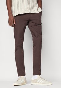 Barna pamut nadrág slim fit szabással, világos bézs sportcipőkkel, amelyek három csíkkal rendelkeznek, és egy textúrált krémszínű inggel.