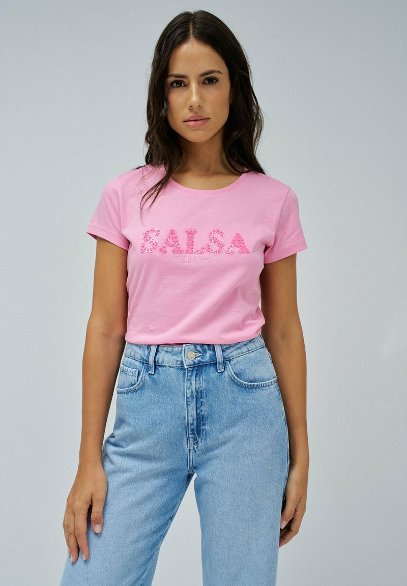 Camiseta de algodón rosa con escote redondo y mangas cortas, que presenta el logo en relieve "SALSA" en un diseño estampado. Se lleva con un denim de color azul claro.