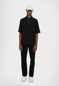 Svart kortermet polo med halv-glidelås og preget logo, kombinert med svarte bukser og hvite joggesko. Kappen er lys blå med kontrasterende tekst.