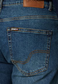 Donkerblauwe denim jeans met een bruine leren label en een klein geborduurd logo op de achterzak. Heeft oranje stiksels en een klassieke uitstraling.