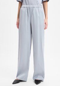 Vrouw in lichtgrijze wijde broek met hoge taille en elastische tailleband, met zwarte puntige ballerina's, op een witte achtergrond.