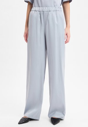 Vrouw in lichtgrijze wijde broek met hoge taille en elastische tailleband, met zwarte puntige ballerina's, op een witte achtergrond.